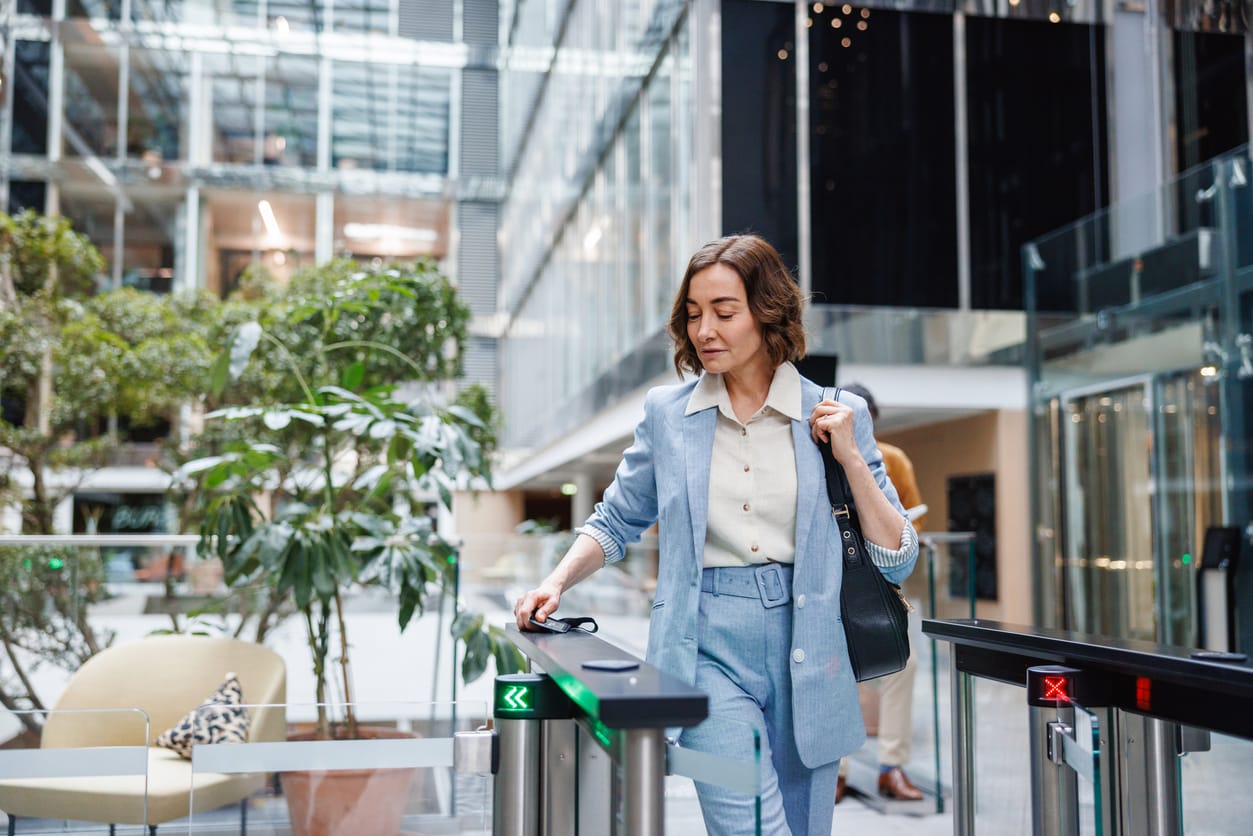 A businesswoman in smart casual attire confidently walks through security turnstiles into a glass-structured office building, ready for the workday.