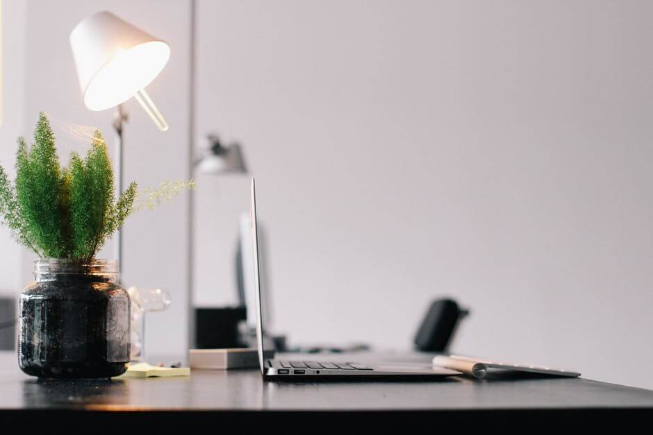 A sleek desk with a laptop, a lamp, and a potted plant in a jar, set against a plain wall background.