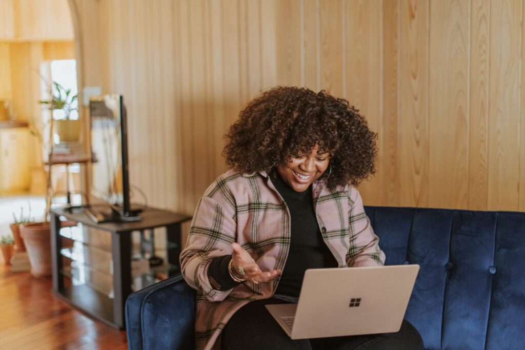 A person with curly hair sits on a blue sofa, smiling and gesturing while looking at a laptop in a cozy room with wooden walls and plants in the background, perhaps engaging in a virtual conversation about generations in the workplace.
