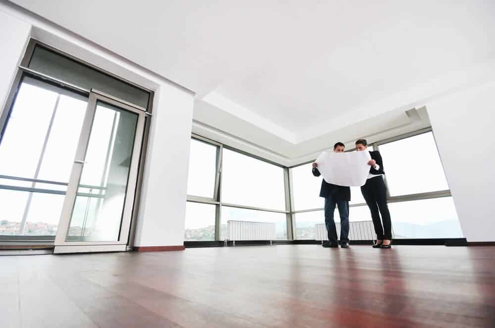 Two people examine a blueprint in a spacious, unfurnished room with large windows and wooden floors.