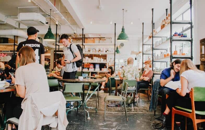 Café scene with people sitting at tables, engaging in conversation and studying, surrounded by shelves and decor.