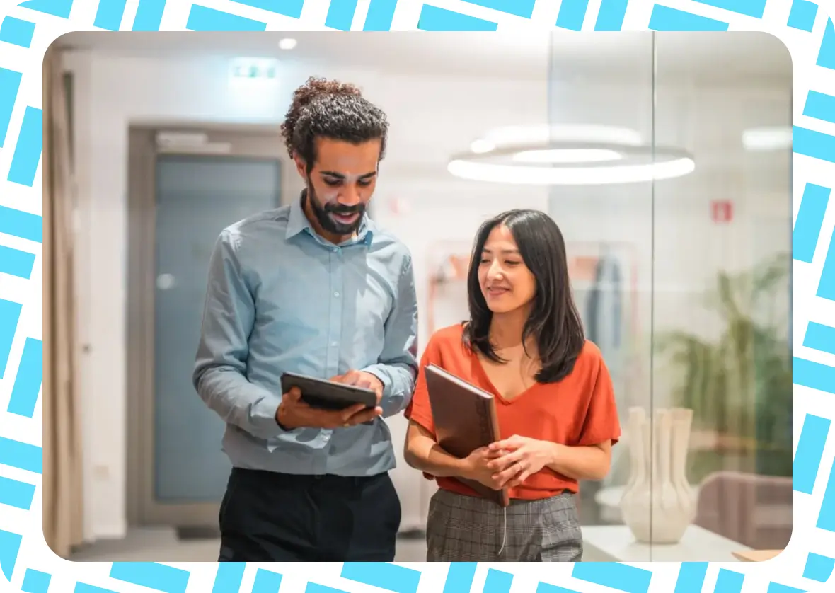 Two people stand in an office, one holding a tablet and the other holding a notebook, smiling and talking. The image has a white and blue patterned border.
