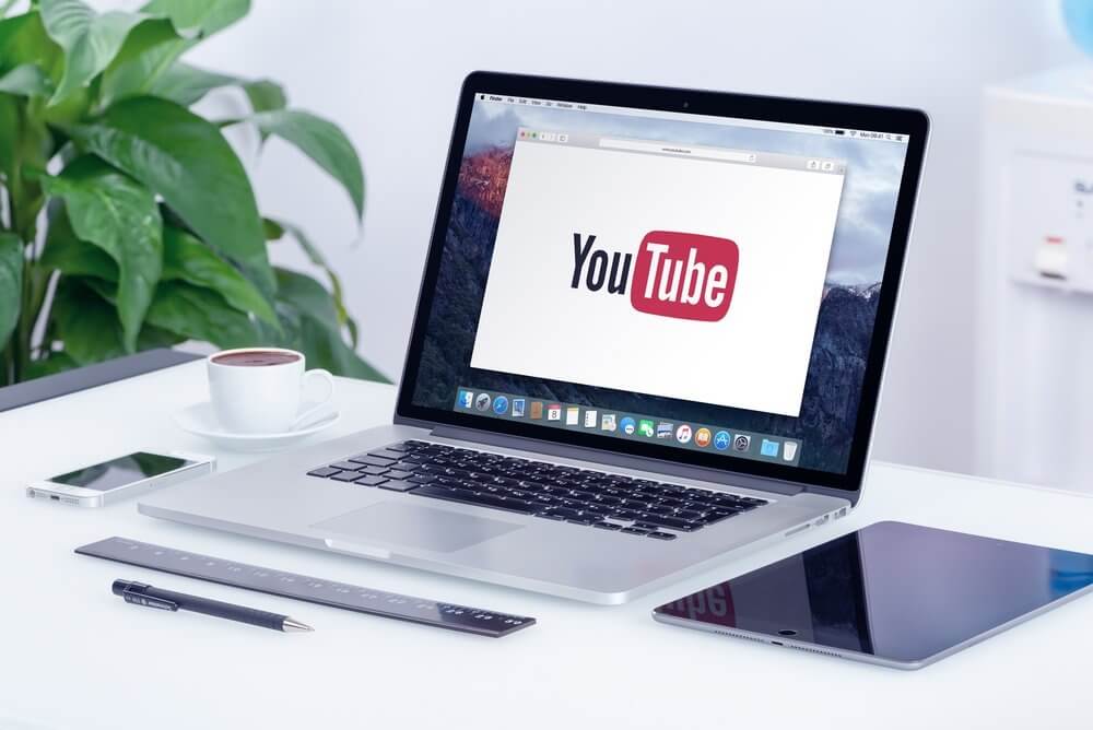 Laptop on a desk displaying the YouTube logo, beside a tablet, ruler, pen, notepad, and cup of coffee.